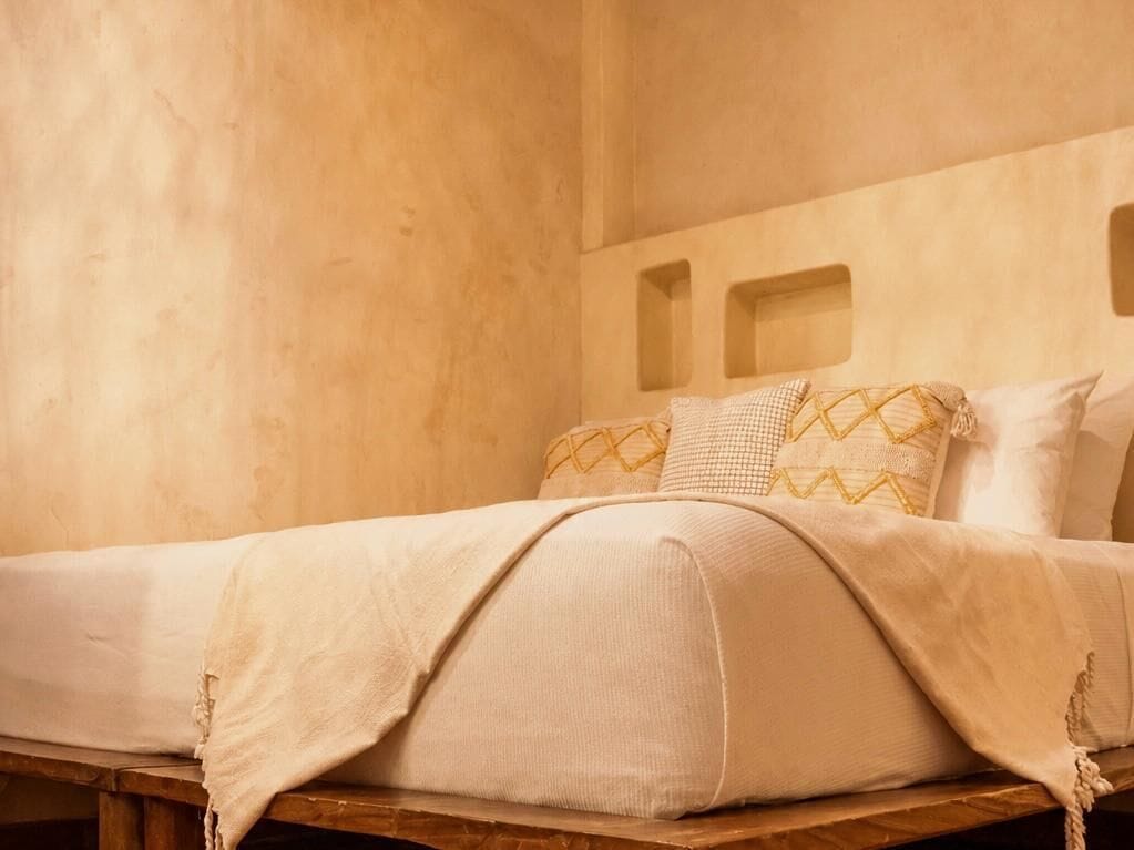 A bed with a beige blanket and pillows, a wooden headboard, and a plain wall.