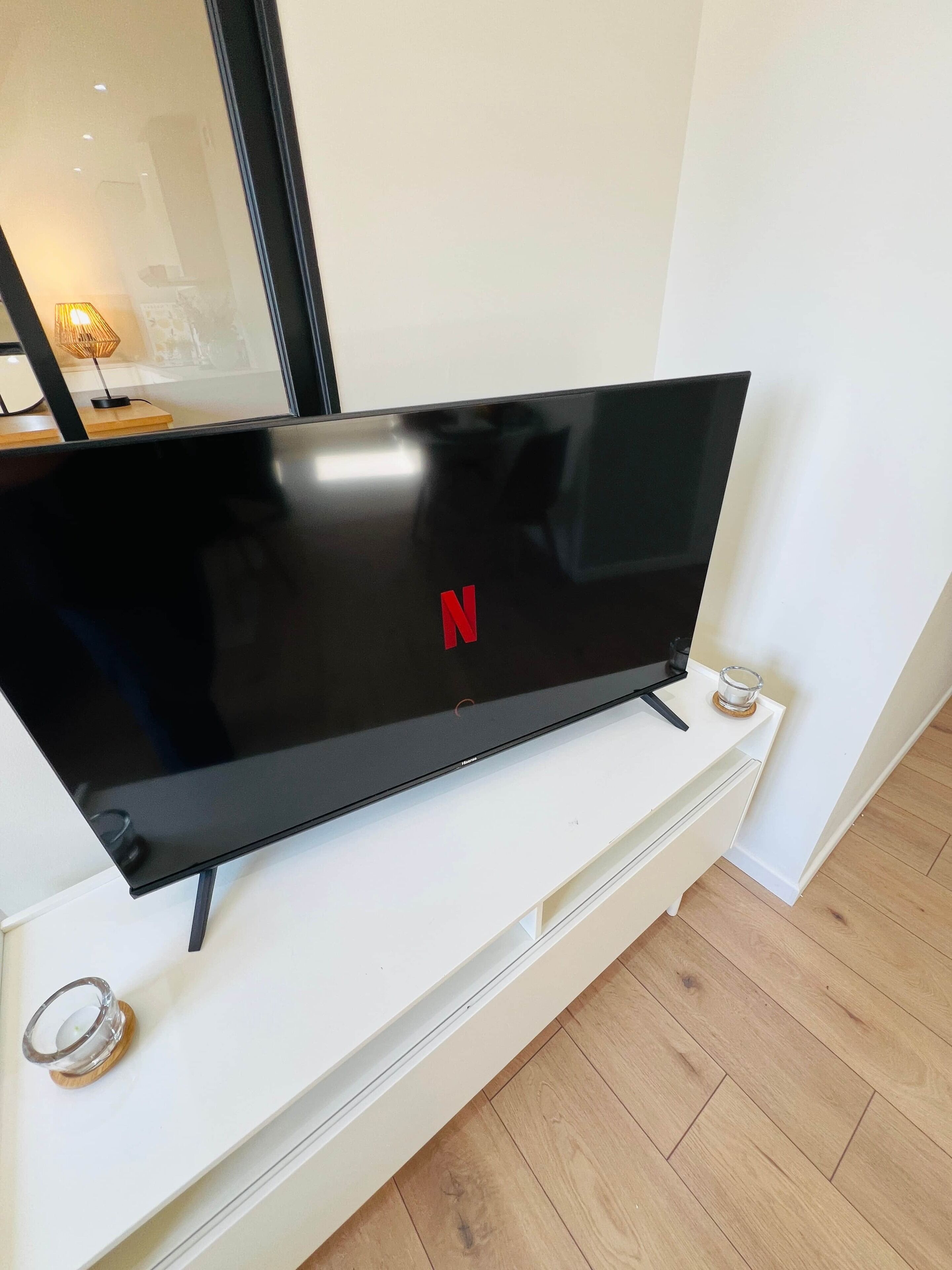 A flat-screen TV on a white stand with a Netflix logo, a glass candle holder, and a wooden floor.