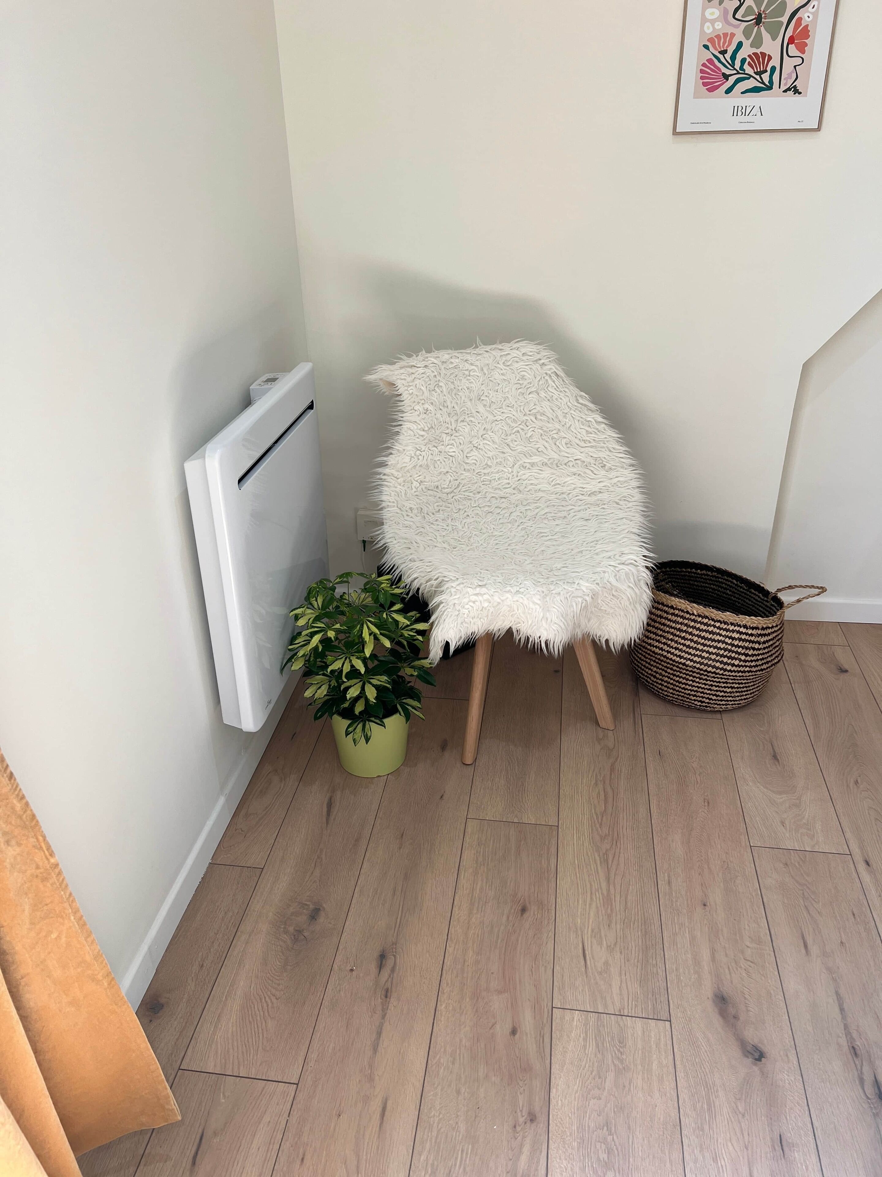 Un coin de pièce avec un radiateur blanc, un fauteuil blanc moelleux, une plante verte en pot et un panier en osier posé sur un plancher en bois.