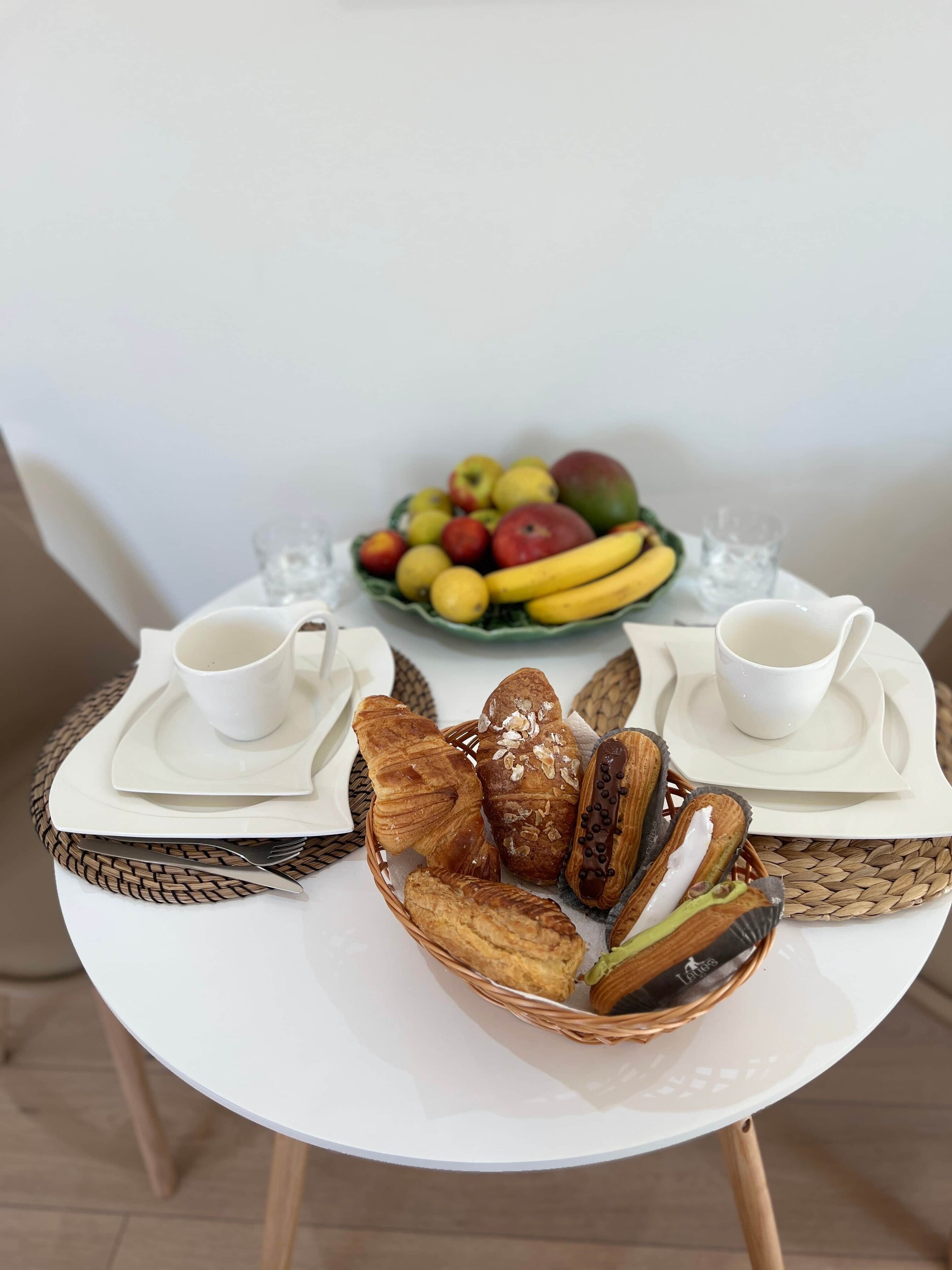 A breakfast setting with a white table, two white cups, a basket of pastries, and a bowl of fruit.