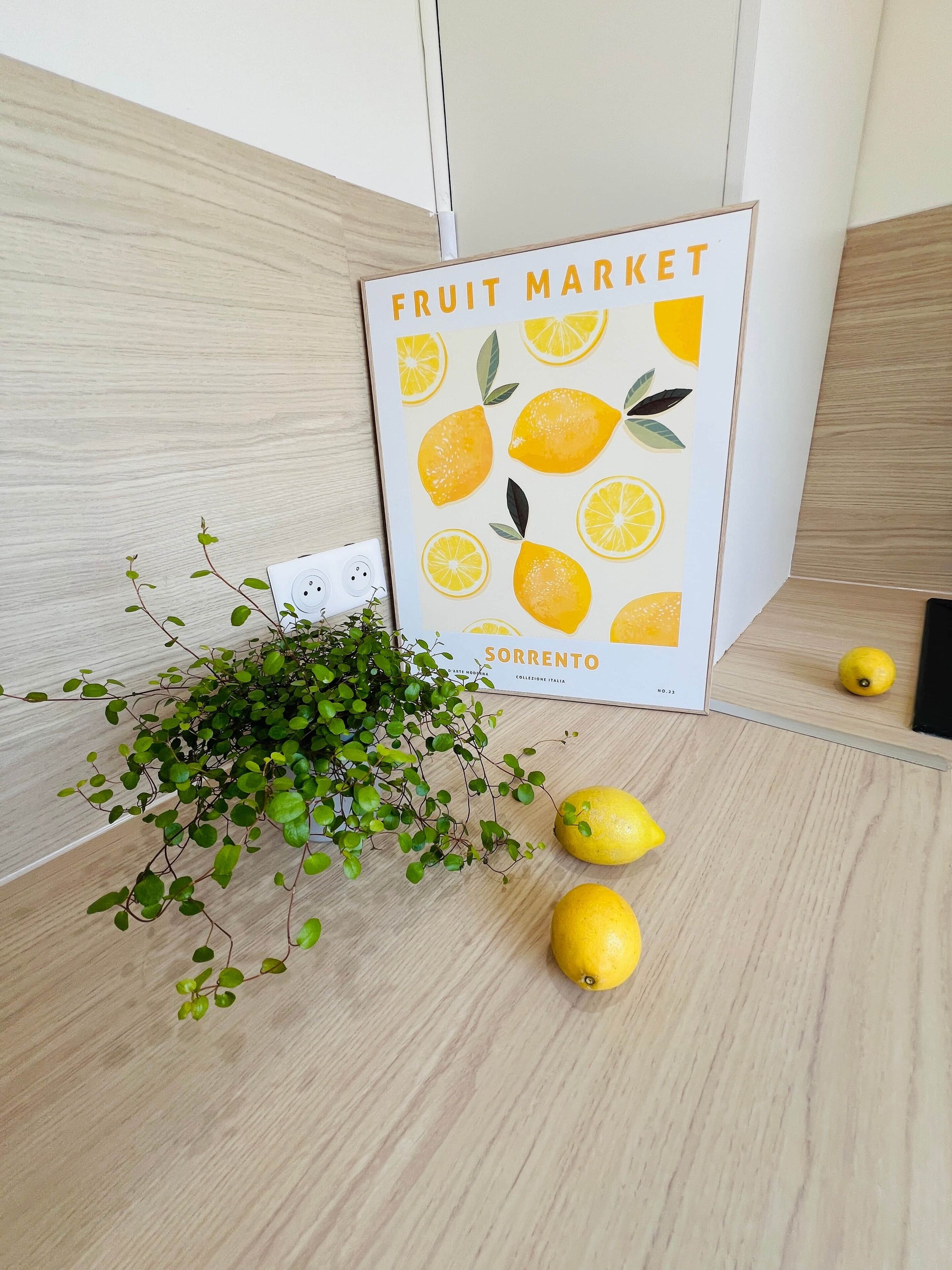 Une affiche sur le thème d’un marché aux fruits, avec une plante en pot et deux citrons posés sur une surface en bois.
