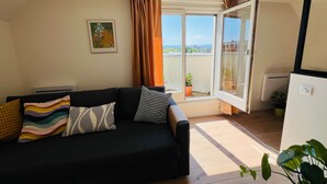 Ein Wohnzimmer mit schwarzem Sofa, gemusterten Kissen, einem gerahmten Bild an der Wand, einem Fenster mit Vorhängen und einer Topfpflanze.