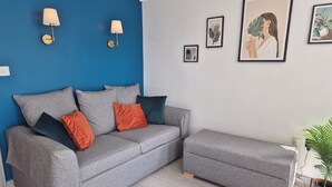 Um sofá cinzento com almofadas laranja e cinzento escuro, um banquinho a combinar e duas luminárias fixadas na parede.
