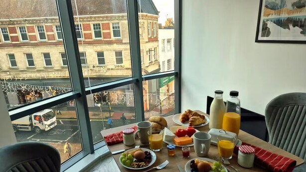 Mesa de café da manhã com pratos de comida, bebidas e vista para prédios através de grandes janelas.