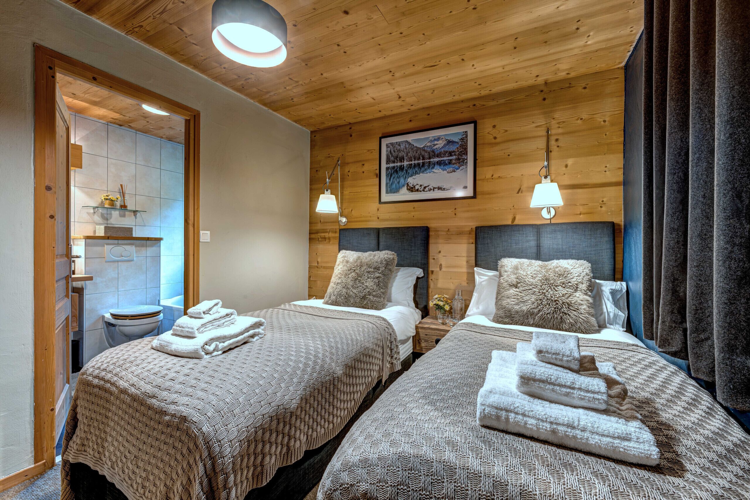 Ein Hotelzimmer mit zwei Betten, Holzboden, einem Bild und einem durchgegangenen Tür sichtbar werdenden Badezimmer.