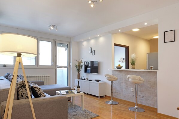 A modern living room with a sofa, a coffee table, and a lamp. There is a kitchen area with a countertop and a bar stool. - Belgrade Central Residence (Belgrade)