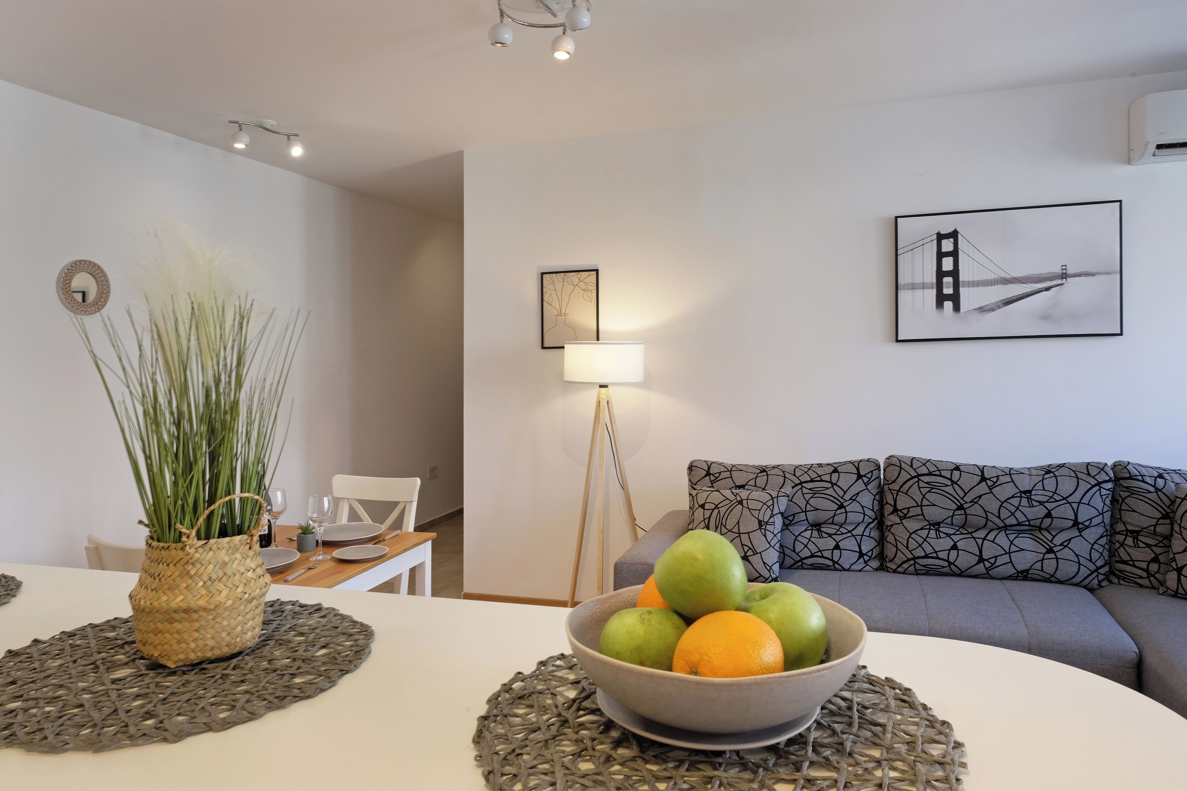 Un salon moderne comprenant un coin repas, un canapé agrémenté de coussins à motifs, un saladier rempli de fruits, une lampe et un tableau encadré représentant un pont.
