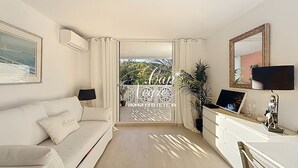 Ett modernt vardagsrum med en vit soffa, en platt-TV, ett skrivbord med en lampa och ett stort fönster med gardiner.