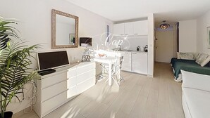 Ett modernt vardagsrum med en vit soffa, ett matbord med stolar, en tv på ett vitt skåp och en stor spegel på väggen.