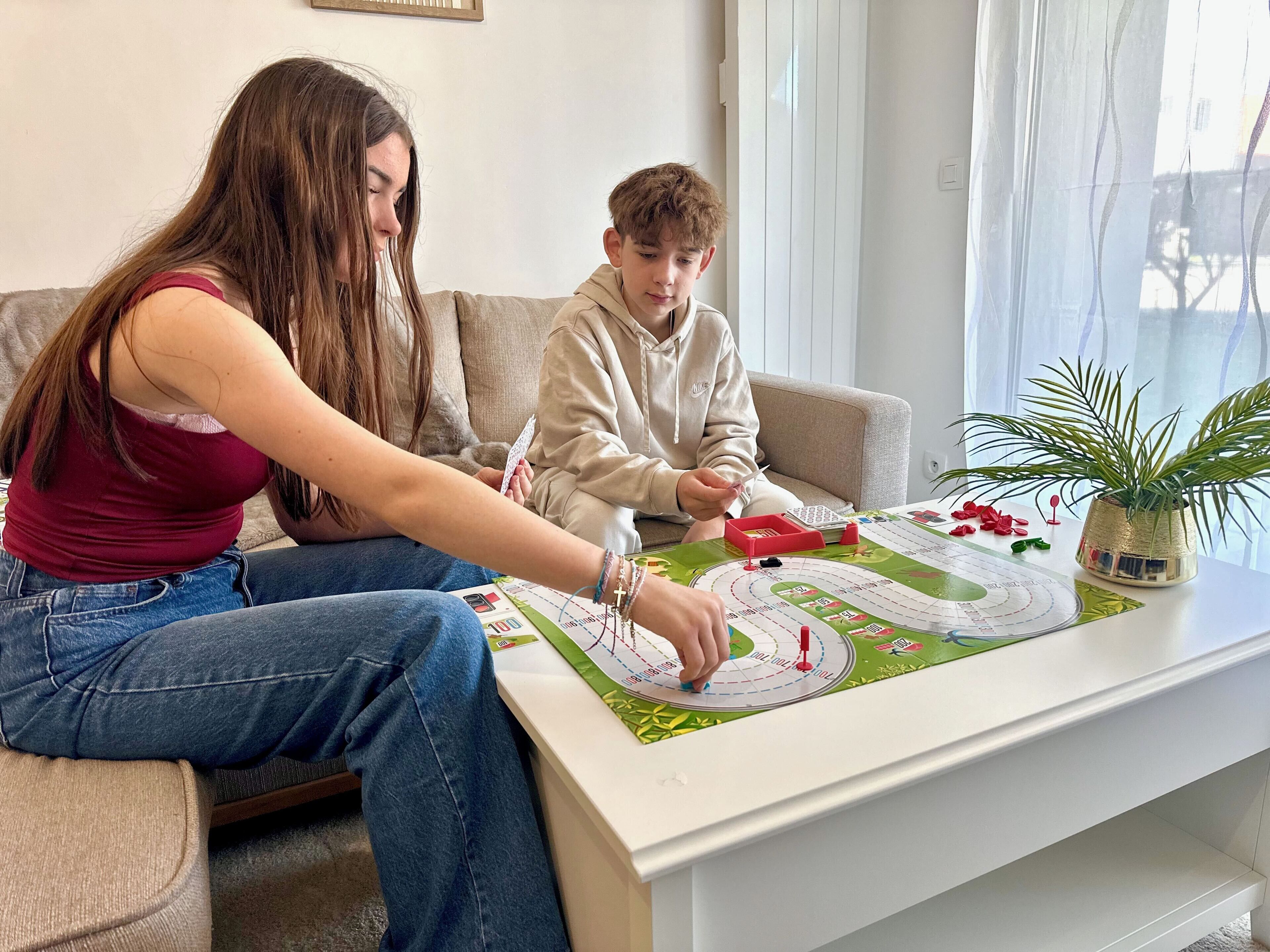 Two people playing a board game on a white table with a green game board, a potted plant, and a window with curtains in the background.