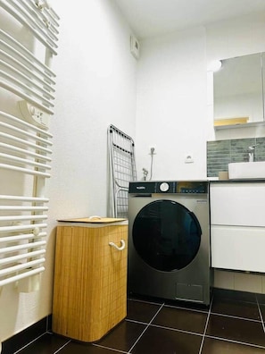 A compact laundry room with a front-loading washing machine, a drying rack, and a small wooden cabinet. - Urban cocoon, quiet studio 2 minutes from Vannes (Plescop)