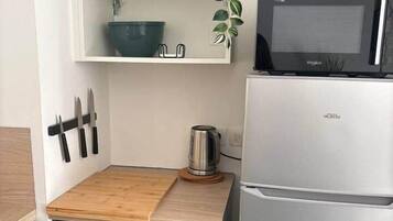 Een kleine keuken met een magnetron, een roestvrijstalen koelkast, een houten aanrecht, een hangplant en een plank met een kom en bestek.