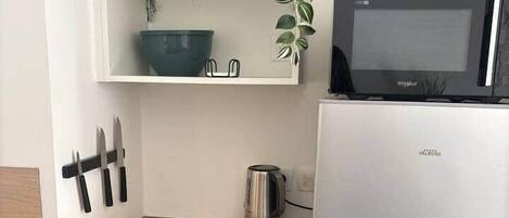 A compact kitchen with a microwave, a stainless steel refrigerator, a wooden countertop, a hanging plant, and a shelf with a bowl and utensils.