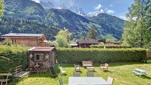 Ein rustikales Holzhäuschen mit Tisch und Stühlen in einer grasbewachsenen Fläche, umgeben von Bergen und Bäumen.