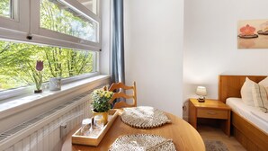 Une pièce comprenant un lit, une table de chevet, une table avec un plateau, une chaise, une fenêtre donnant sur des arbres et un tableau accroché au mur.