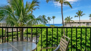 A balcony with a glass table and chair, overlooking a beach with palm trees and a building in the background. - Not available now (Kelowna)