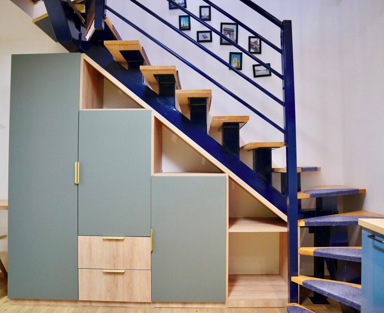 Een trap met een blauwe leuning en houten treden, leidend naar een plateau met een lichtblauwe kast en een houten ladeblok.