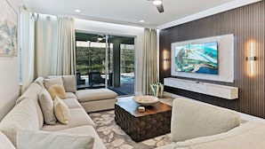 A modern living room with a sectional sofa, a coffee table, a flat-screen TV mounted on a wooden wall, and a ceiling fan. - Magical Memories Villa 10bd Near Disney (Kissimmee)
