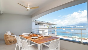 A balcony with a wooden table set for two, white chairs, and a wicker chair. A ceiling fan is visible above the table. The balcony overlooks a marina with boats and a clear blue sky. - Odyssée Bleue (Trois-Ilets)