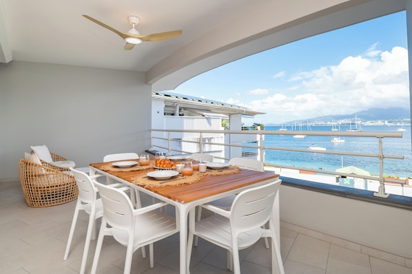 A balcony with a wooden table set for two, white chairs, and a wicker chair. A ceiling fan is visible above the table. The balcony overlooks a marina with boats and a clear blue sky. - Odyssée Bleue (Trois-Ilets)