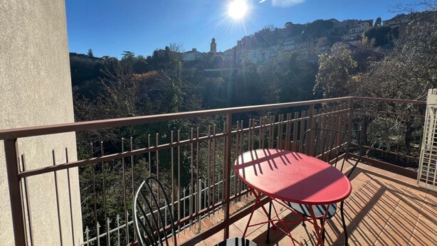 Ein Balkon mit einem roten Tisch und Stühlen, einem Metallgeländer und Blick auf eine Hügelkuppe mit Gebäuden.