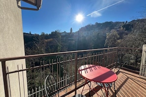 Un balcón con una mesa y sillas rojas, baranda de metal y vista a una colina con edificios.