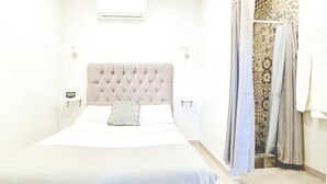 A neatly made bed with a tufted headboard, a bedside table with a clock, and a shower area with patterned tiles. - PrivateStudio/DoubleBed+CouchBed/Near Border (Tijuana)