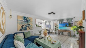 A living room with a blue sofa, a green table, and a TV displaying a beach scene. - Garage, Gated Yard, near SD Gaslamp, DT, 4BR/2Bth (National City)