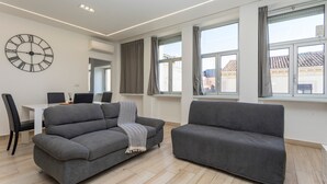Un saló modern amb un sofà gris, una taula de dinar amb caires i un rellotge de paret gran.