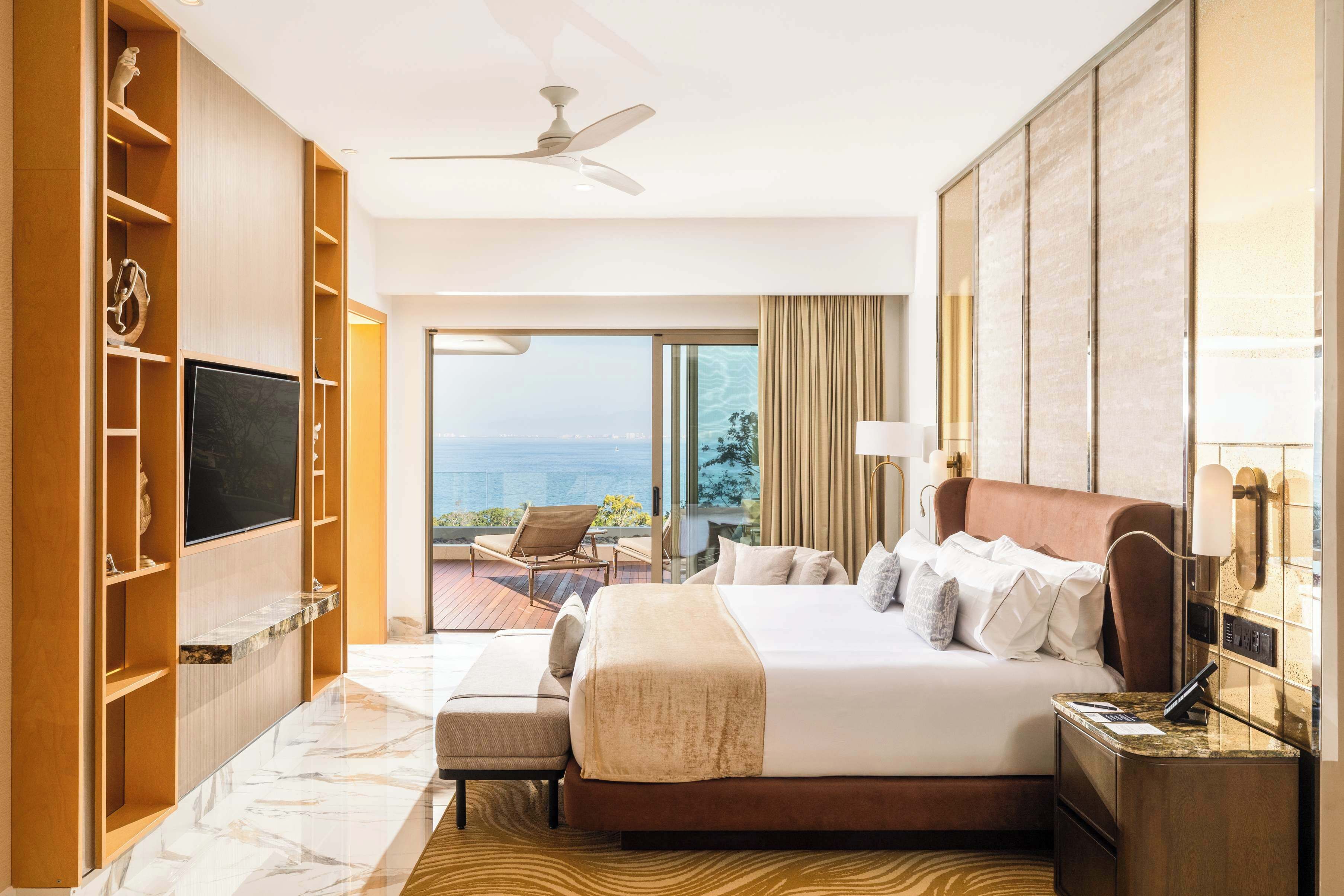 Quarto de hotel com cama, televisão, varanda com cadeira e vista para o mar.