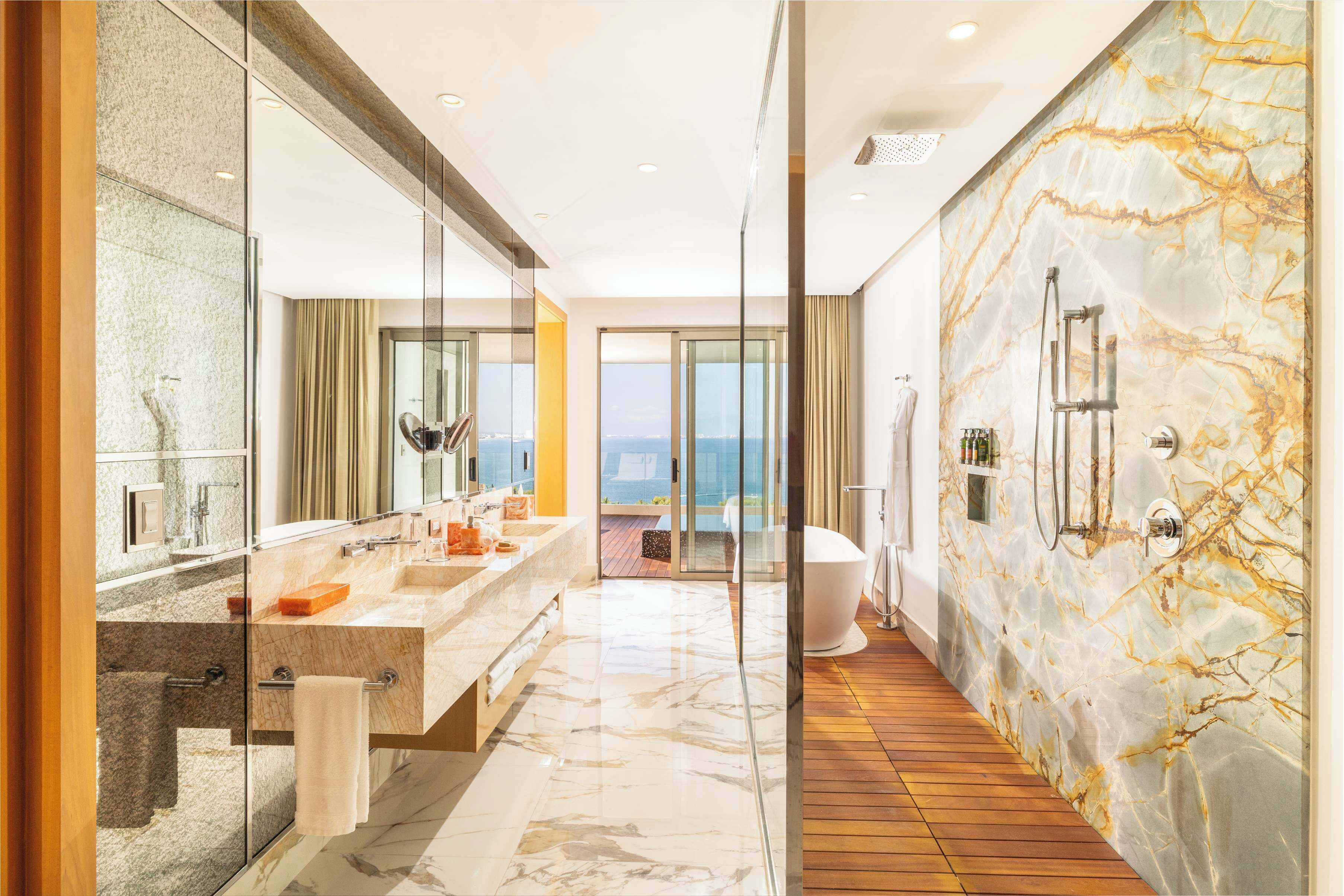 A modern bathroom with a large mirror, two sinks, a bathtub, and a view of the sea.