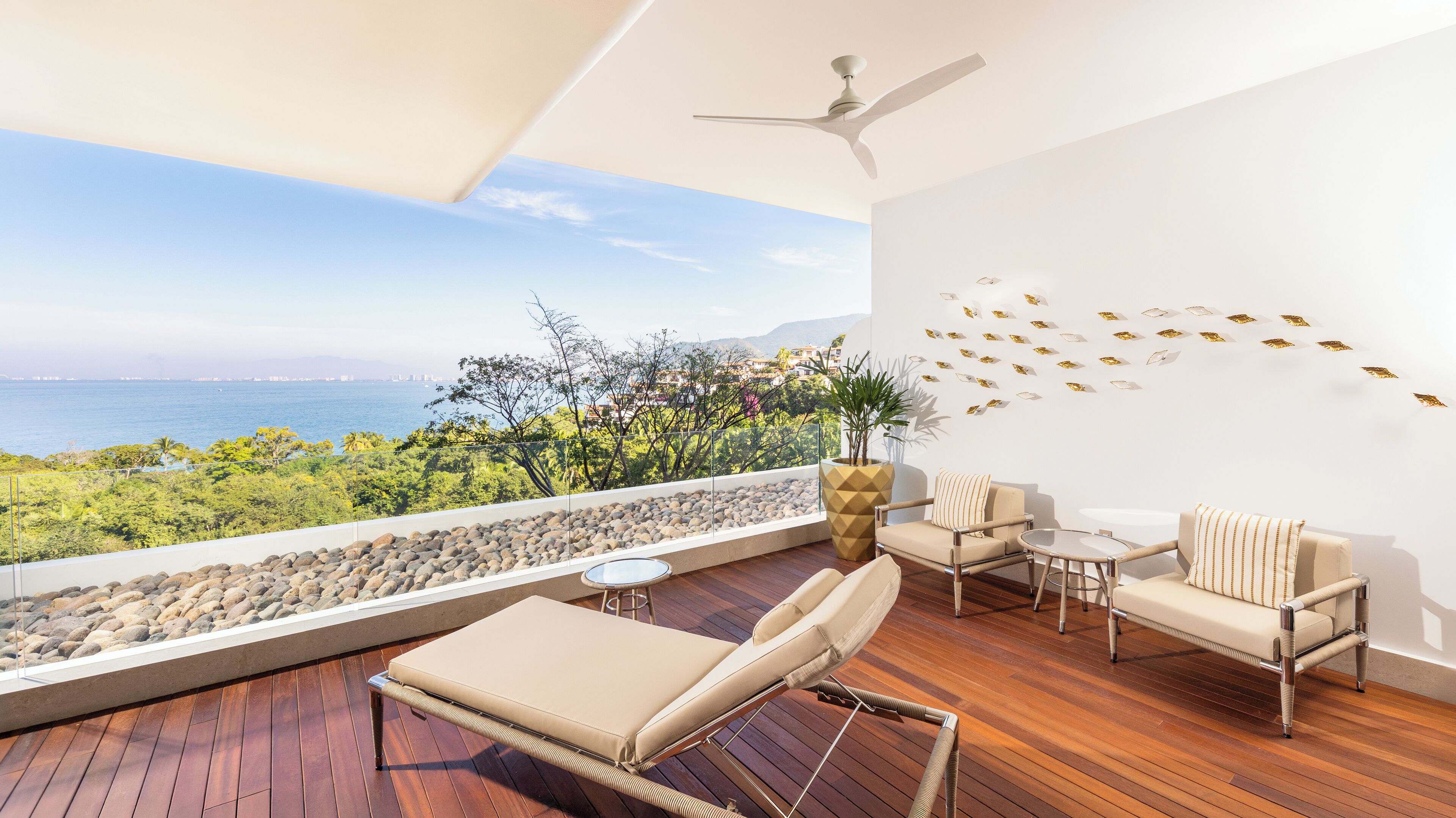 A balcony with a view of the sea, furnished with a lounge chair, two armchairs, and a small table.