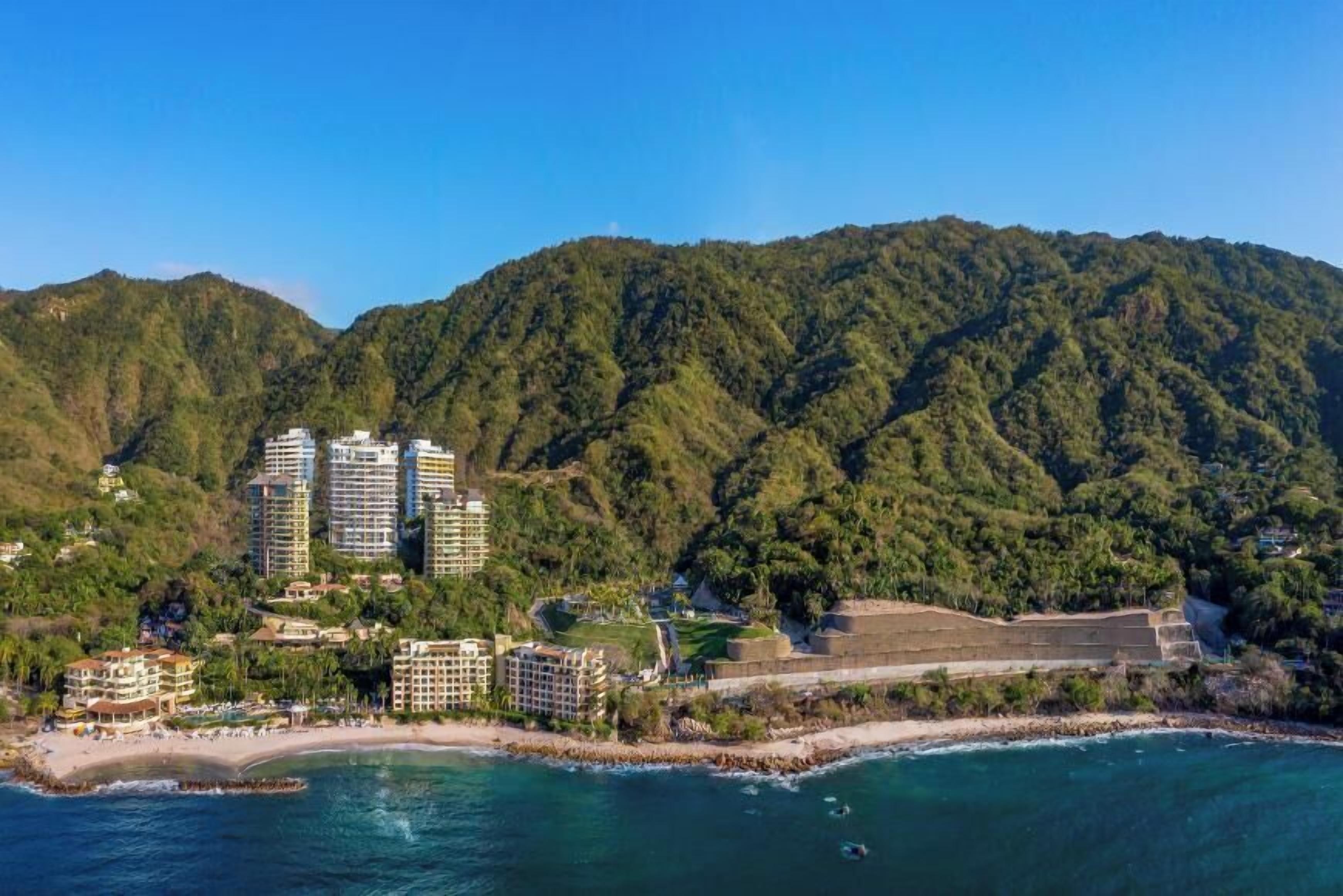 Un resort costero con villas frente a la playa, un campo de golf y montañas al fondo.