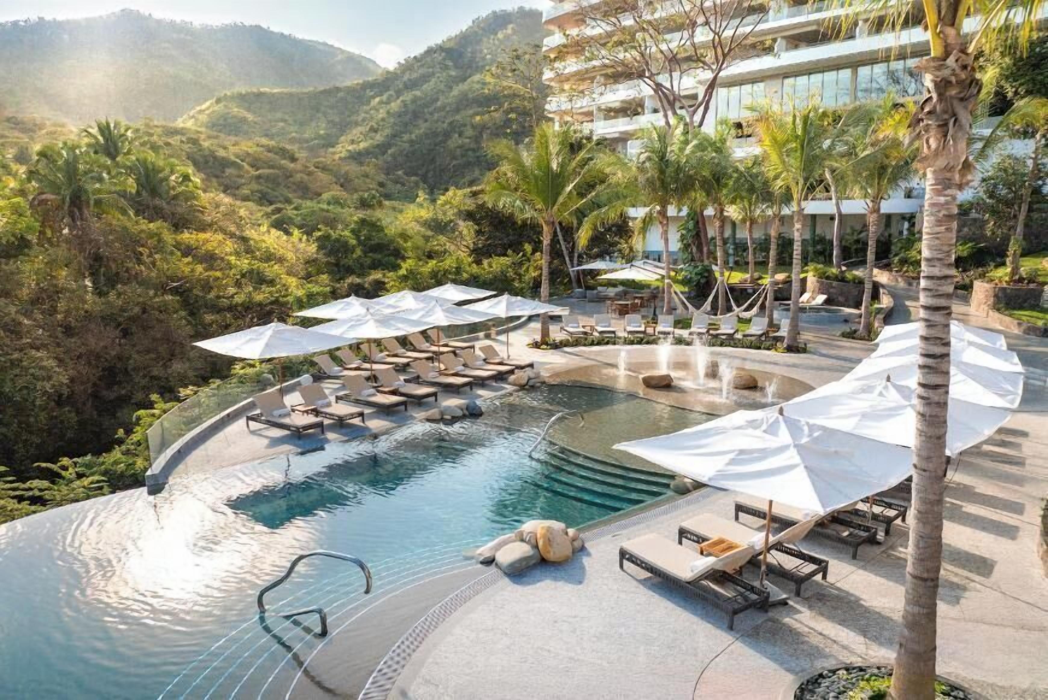 A pool area with lounge chairs and umbrellas, surrounded by palm trees and a mountainous backdrop.