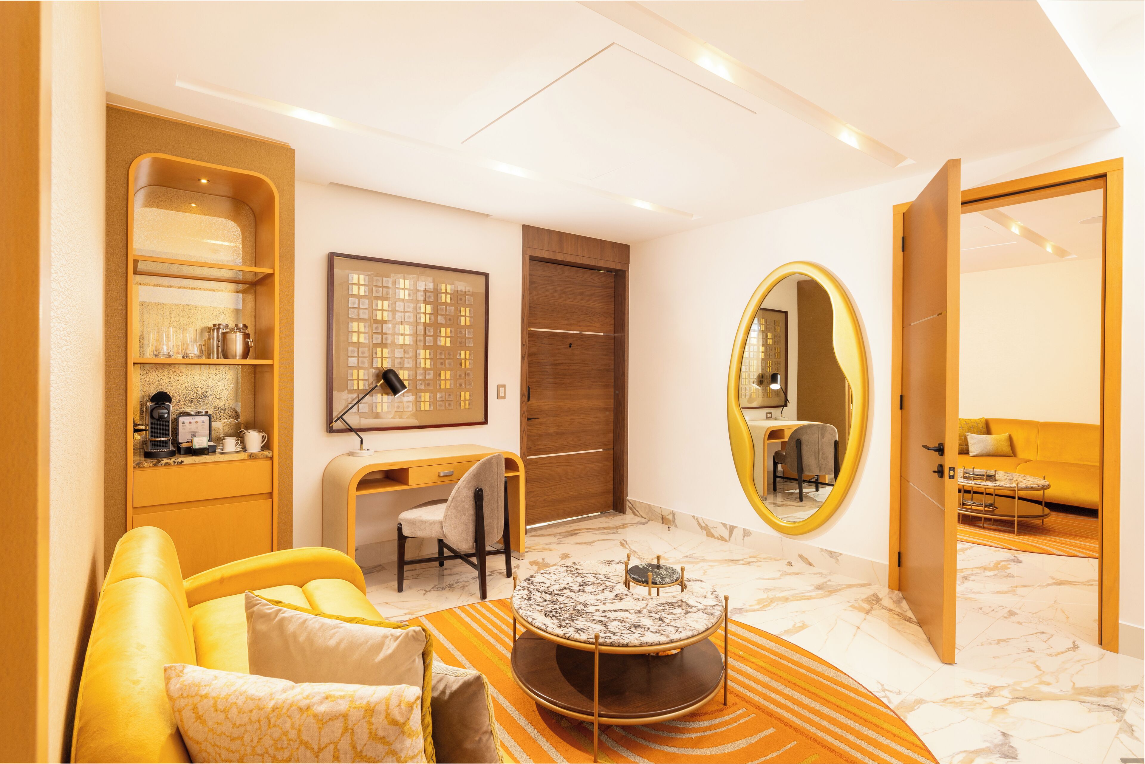 A modern hotel room with a yellow sofa, a round coffee table, a desk with a chair, a large mirror, and a wooden wardrobe.