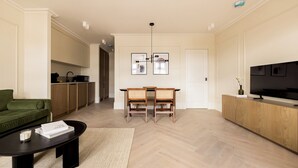 Una sala de estar moderna con un sofá verde, una mesa de centro, una mesa de comedor con sillas, un televisor sobre un mueble de madera y una zona de cocina al fondo.