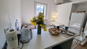 Pieni, käytännöllinen keittiö, jossa on valkoinen työtaso, vedenkeitin, vesipullo, keltainen kukka-asetelma maljakossa, lautasella ruokaa ja jääkaappi.