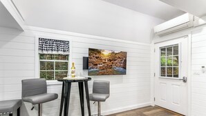 Una habitación con una pared blanca con paneles, un televisor montado, dos taburetes grises, una pequeña mesa redonda con una botella y un vaso, y una puerta con ventana.