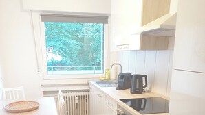 Kompakti keittiö, jossa on valkoiset kaapit, sisäänrakennettu uuni, kahvinkeitin ja ikkuna, josta avautuu näkymä puita kohti.