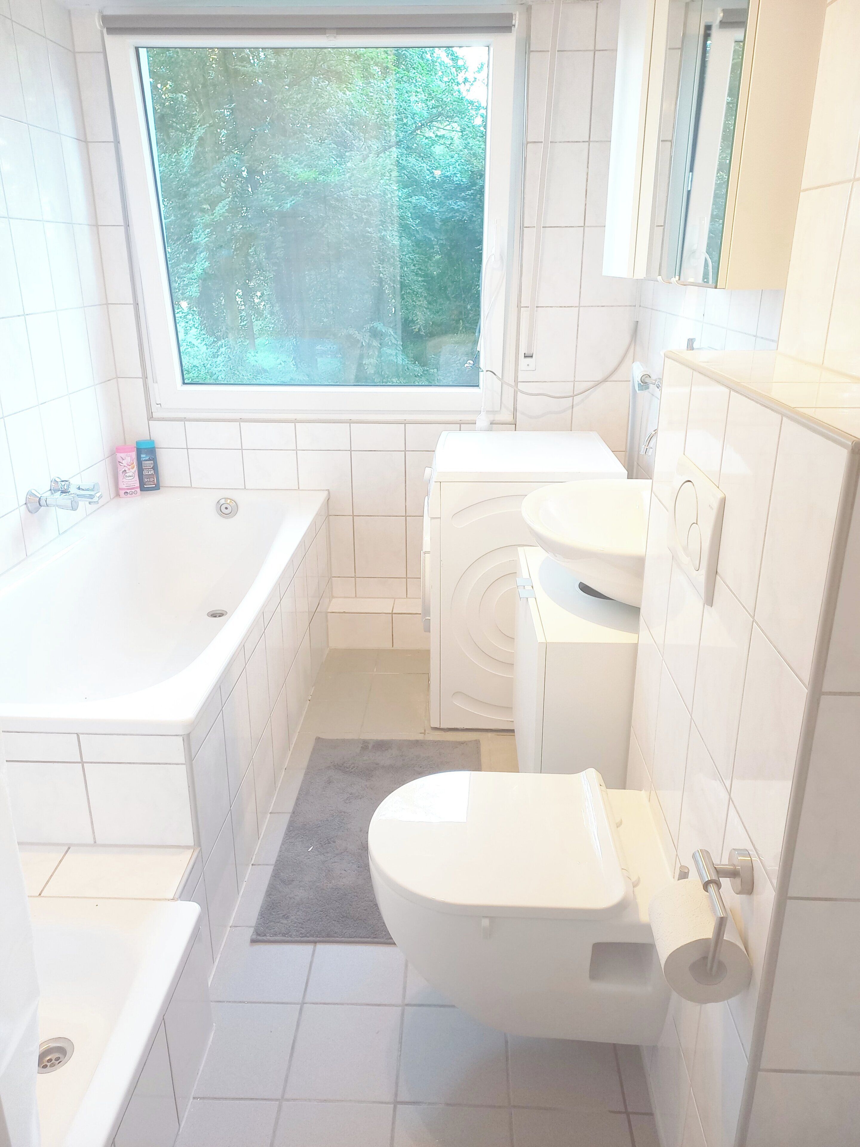 A modern bathroom with a bathtub, toilet, and sink, featuring white tiles and a window with a view of greenery.