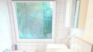 Moderni kylpyhuone, jossa on kylpyamme, wc ja pesuallas. Kylpyhuoneessa on valkoiset laatat ja ikkuna, josta avautuu näkymä vehreään maisemaan.