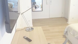 Hotellihuone, jossa on sänky, työpöytä lampun kanssa, tuoli, seinälle asennettu televisio ja ikkuna, jossa on verhot.