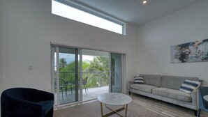 A modern living room with a sofa, a round coffee table, and a chair. There is a sliding glass door leading to a balcony with outdoor furniture.