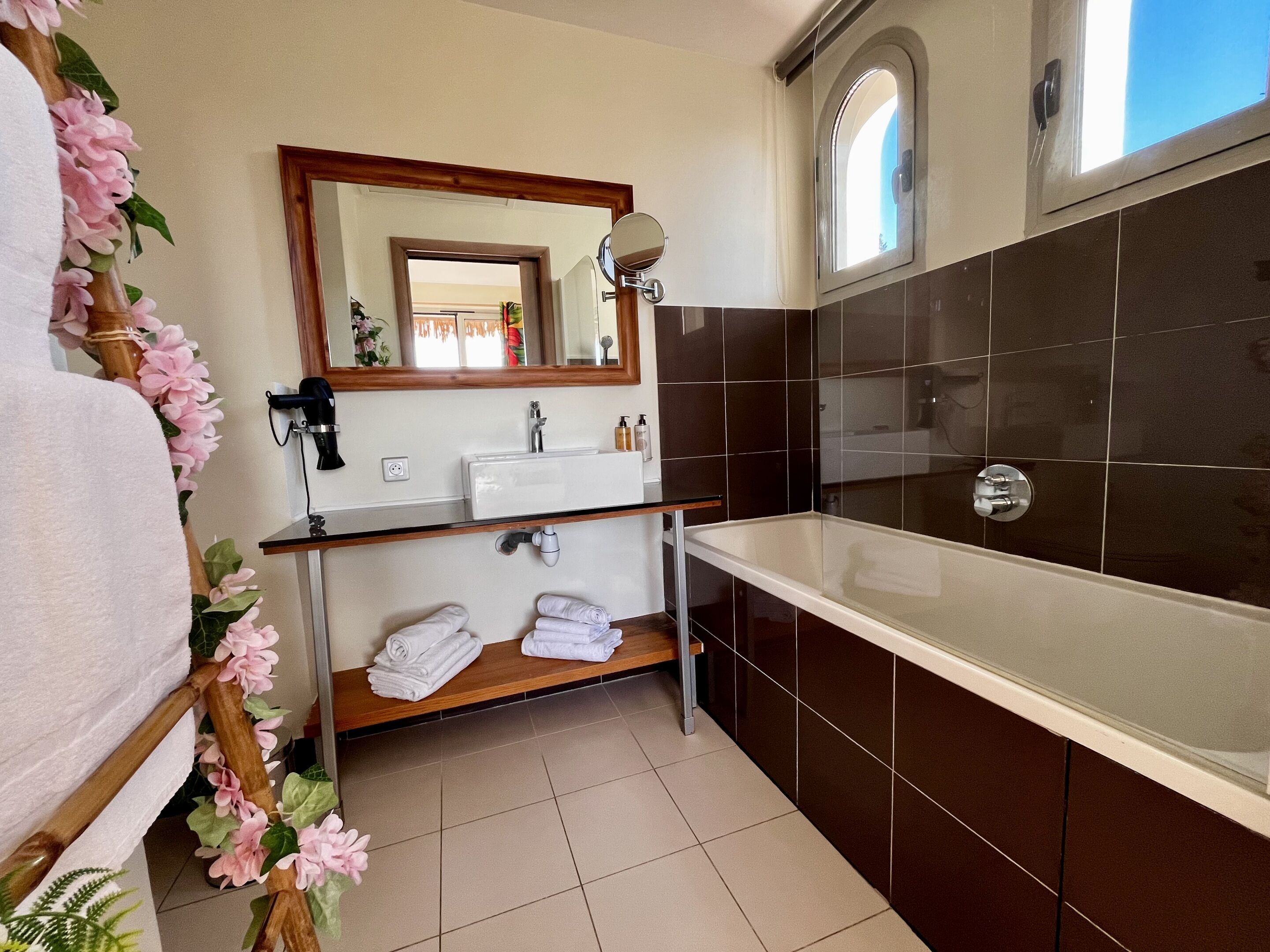 Een moderne badkamer met een groot bad, een wastafel en een spiegel. Handdoeken liggen netjes opgevouwen op een plank, en de muren zijn betegeld.