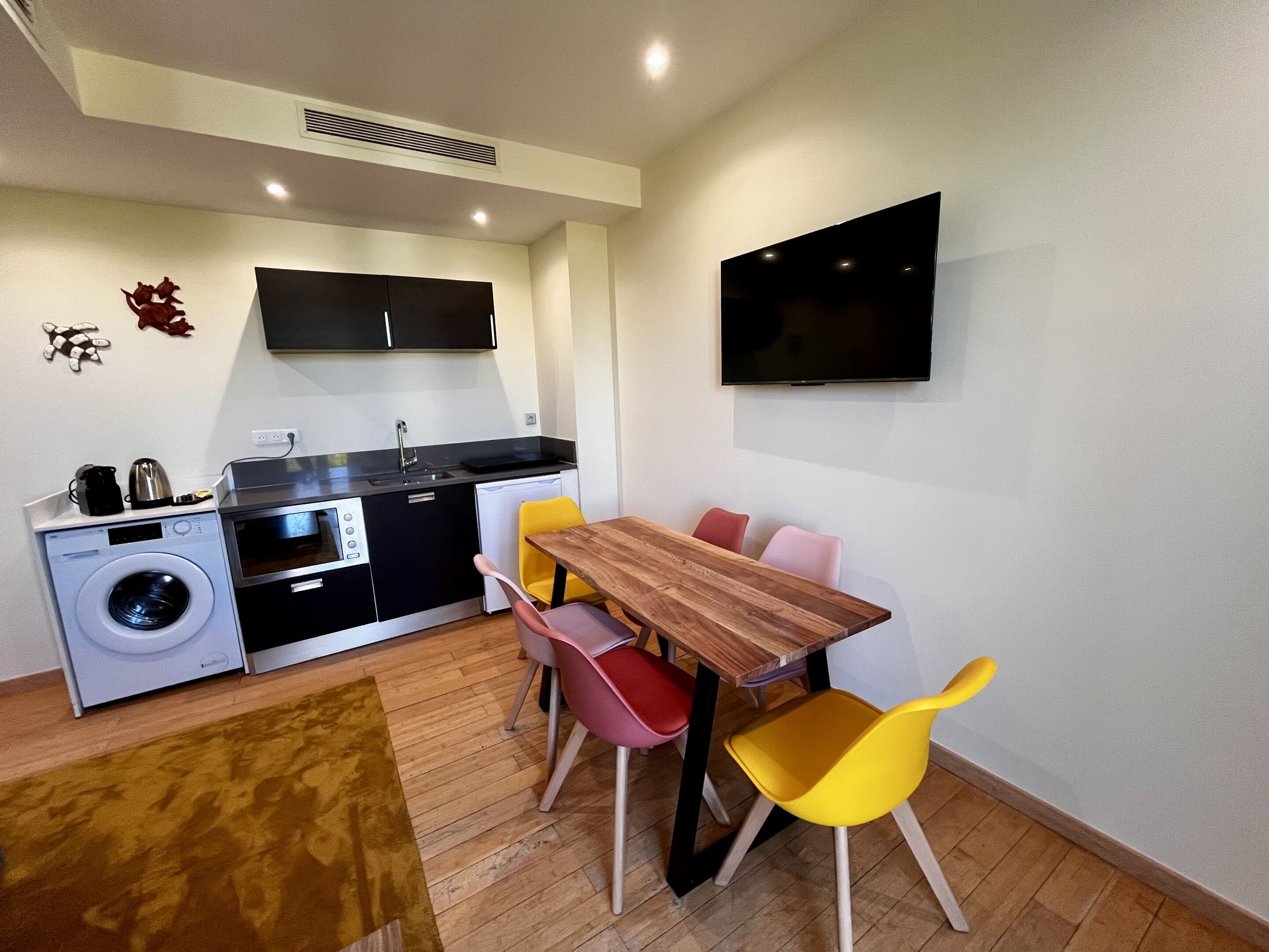 Une cuisine compacte équipée d’un lave-linge, d’un évier et d’un four à micro-ondes. On y trouve une table à manger avec des chaises, ainsi qu’un téléviseur à écran plat fixé au mur.