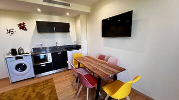 Une cuisine compacte équipée d’un lave-linge, d’un évier et d’un four à micro-ondes. On y trouve une table à manger avec des chaises, ainsi qu’un téléviseur à écran plat fixé au mur.