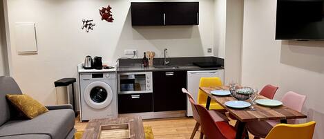 A compact living space with a sofa, dining table, chairs, a washing machine, a small kitchen area, and a TV mounted on the wall.