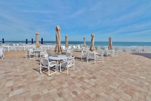 Un área de estar al aire libre con sillas y mesas blancas, sombrillas beige y vista al océano.