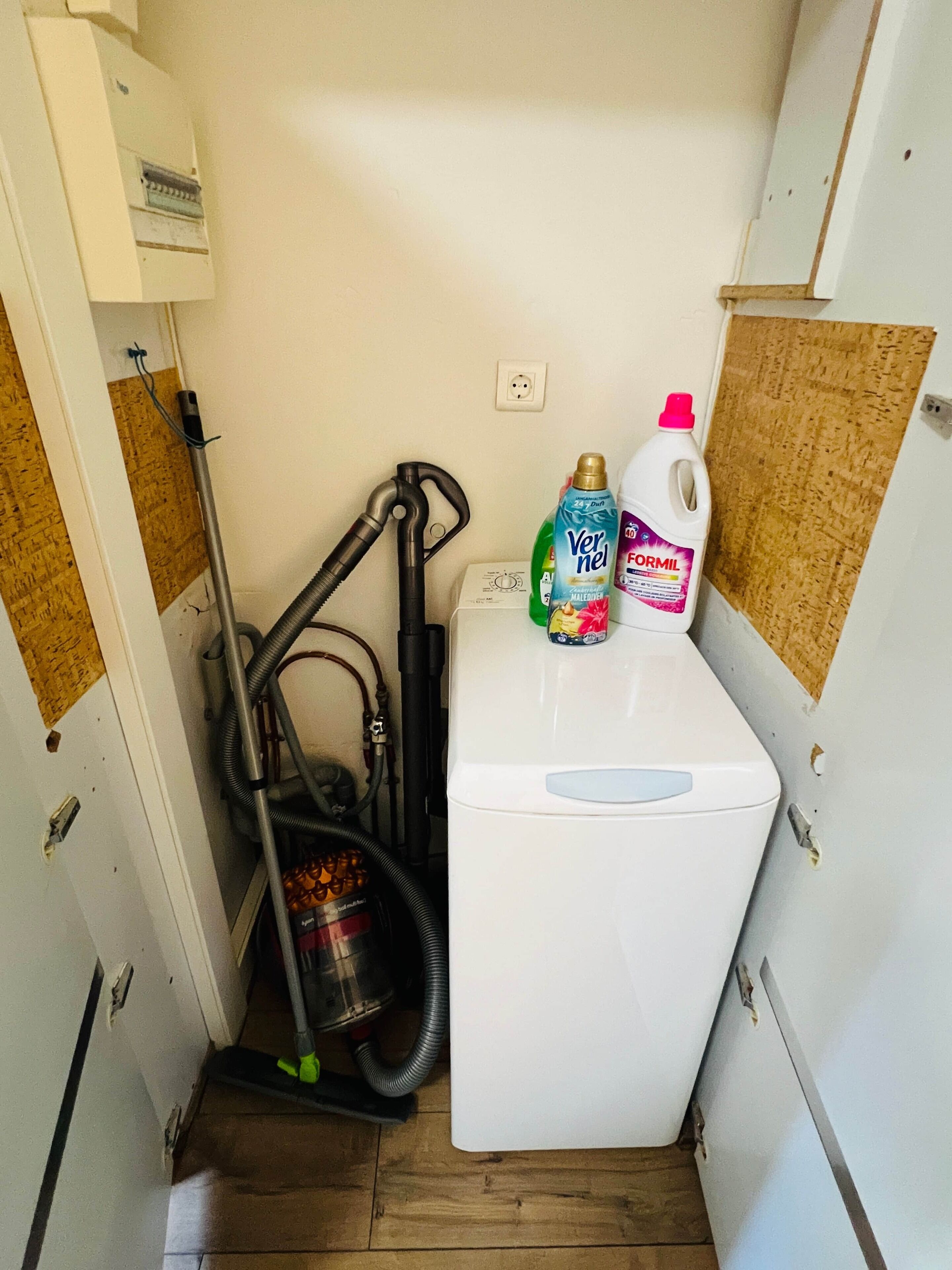 A compact room with a white washing machine, a vacuum cleaner, and cleaning supplies on a small table.