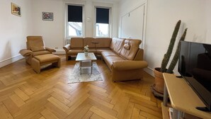 A living room with a sofa, armchair, and coffee table. There are two windows with blinds, a cactus, and a television. - Spacious 160 sqm apartment in central Göppingen for families, fitters, groups (Göppingen)